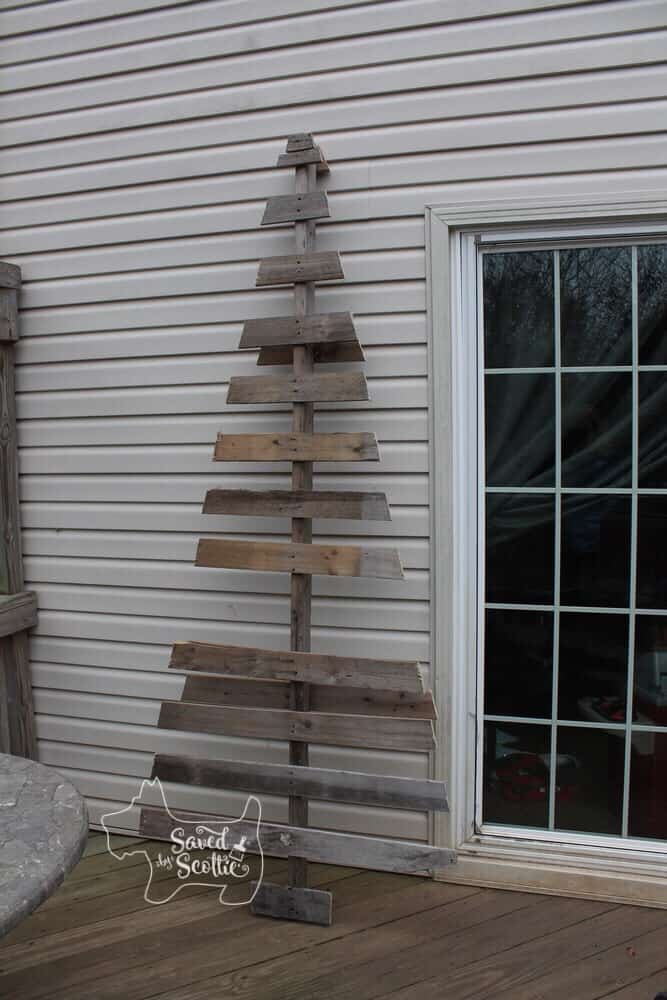 a simple wood pallet christmas tree missing slats leaned against the siding of a house next to a glass door