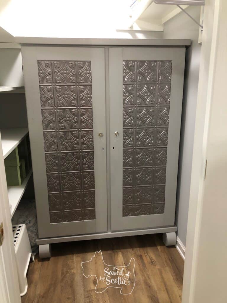 closed armoire with silver metal decorative fasäde panels, glass knobs and in a closet with wood floor and shelves to the left of the armoire. 