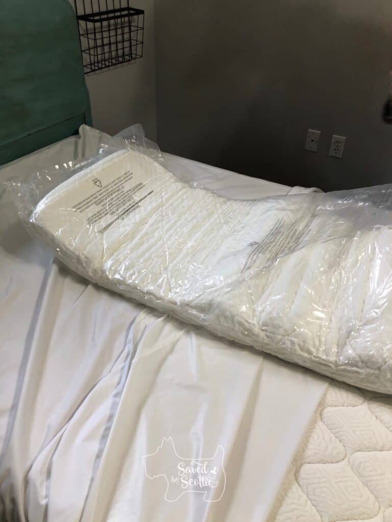 Therapedic memory foam mattress topper in package being unrolled on top of mattress. Bed headboard is teal and wall in background is grey with a basket hanging on the wall.