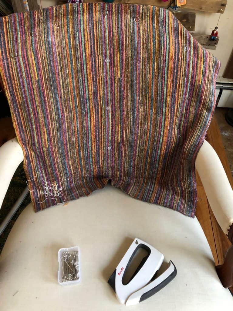 striped fabric draped over chair back and attached with both skewers around the outside edes and twist pins in the center to keep it in place while stapling or tacking in place. Staple gun and box of skewers on seat of chair. 