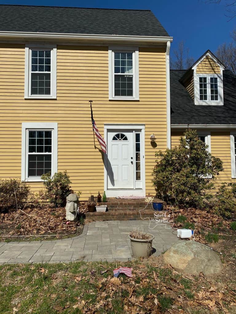 yellow house with white windows and trim. front door is also white with US flag hanging next to it. Concrete easter island head in the front garden an a brick porch with grey stone walkway. 