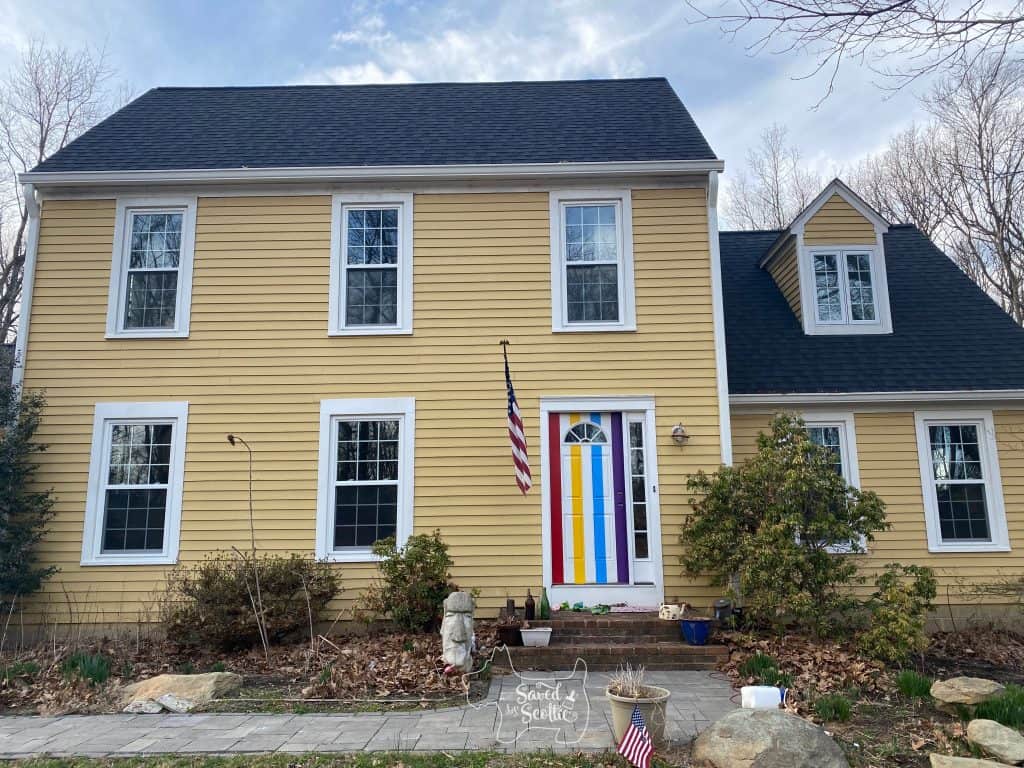 Full view of yellow house with painted front door in progress. stripes of red, yellow, blue, and violet look more like a circus tent at this stage. US flag hanging next to door, easter island head in garden. 