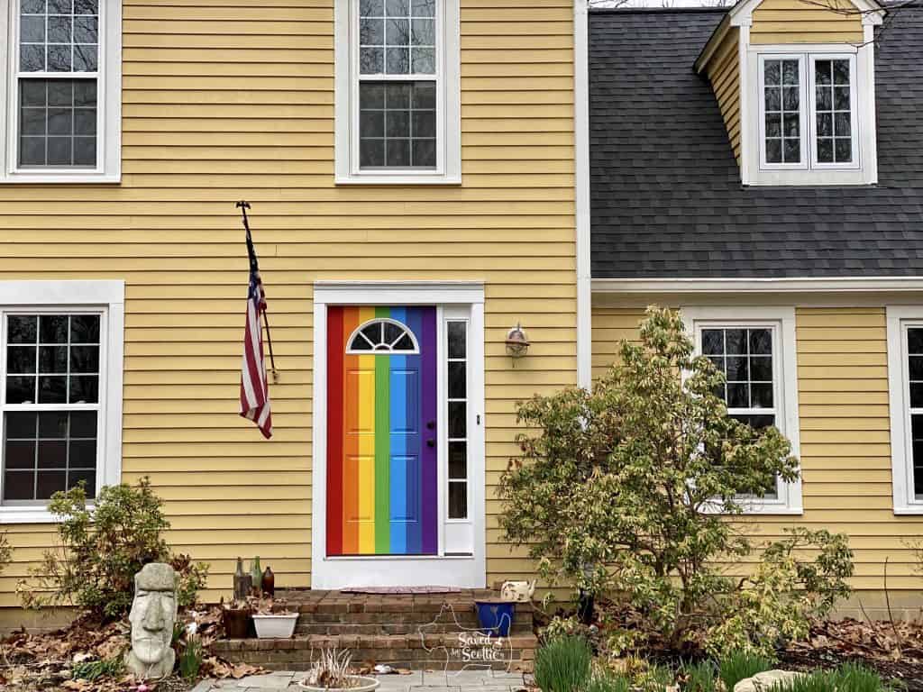 full view of yellow house with rainbow painted door. US flag hanging on left, concrete easter island head in garden, brick landing and stone walkway. 